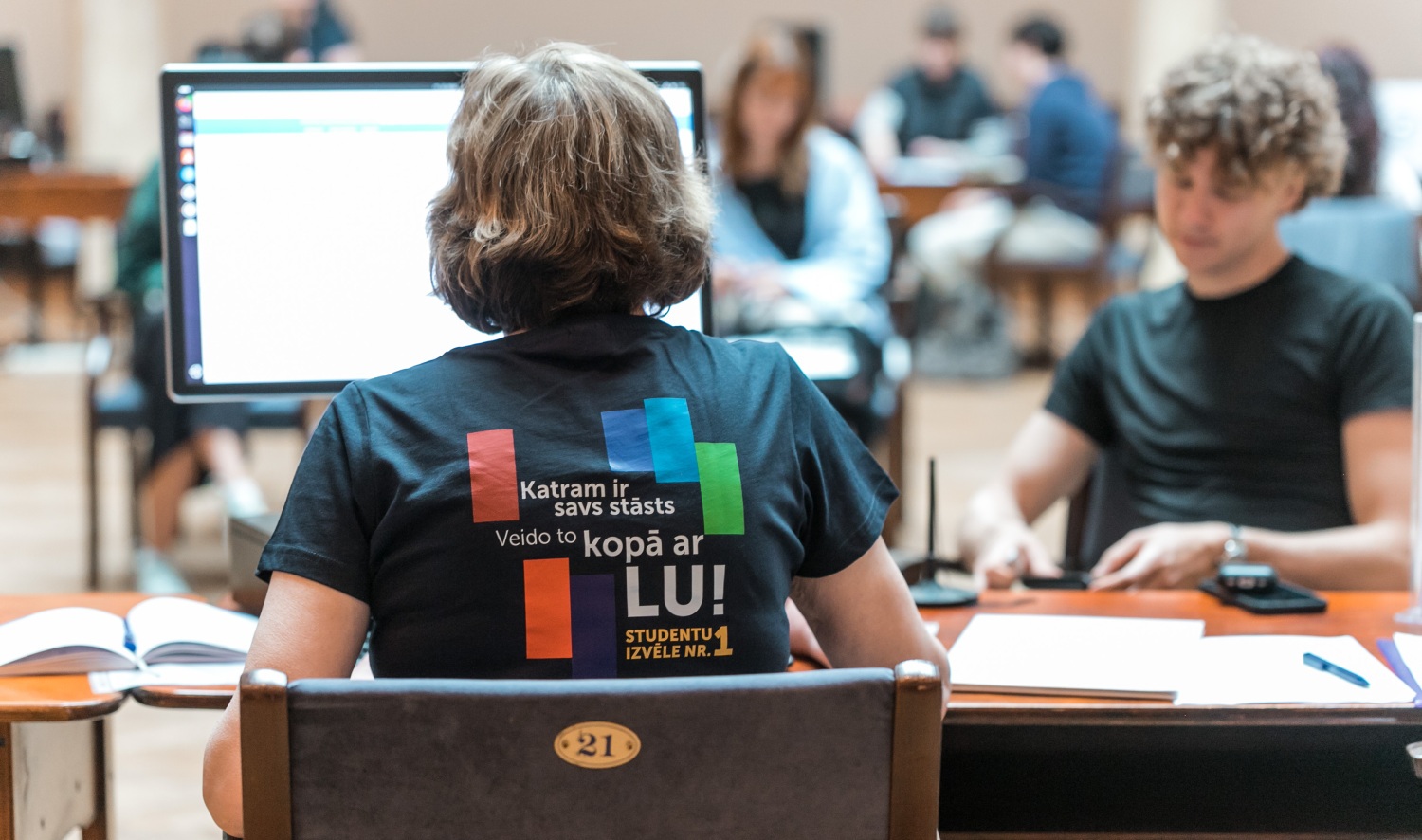 Foto no uzņemšanas. Studētgribētājs sēž pie galda. Studentu servisa darbinieks pieņem viņa dokumentus.