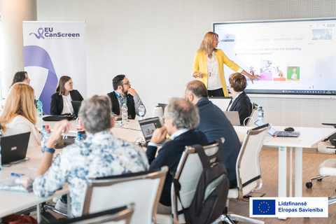 Konferences telpā notiek EU CanScreen projekta sanāksme: cilvēki sēž pie galda ar klēpjdatoriem, bet sieviete pie ekrāna prezentē informāciju. Attēla labajā apakšējā stūrī redzams Eiropas Savienības līdzfinansējuma logotips