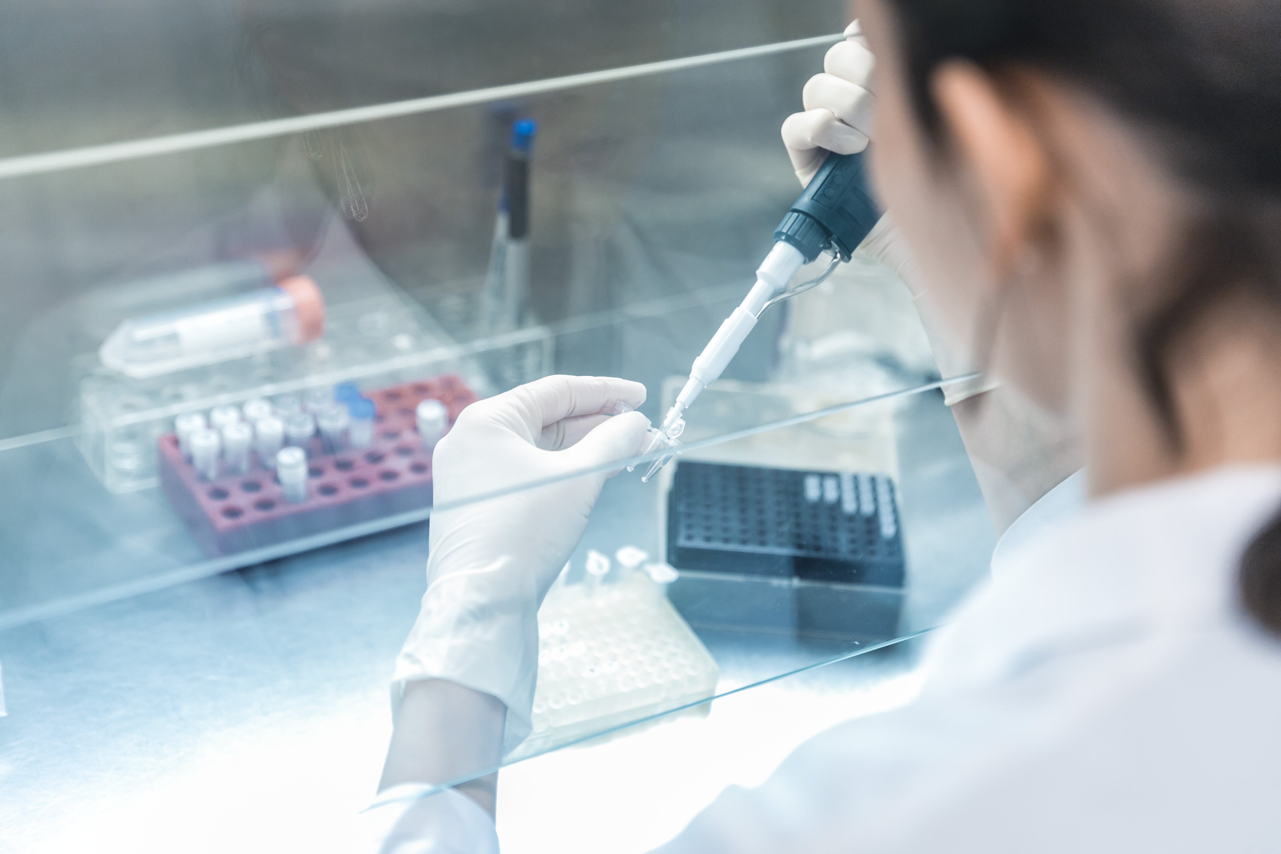 Laboratorijas darbinieks ar baltām cimdiem izmanto pipeti, lai pārvietotu šķidrumu mazā caurulītē sterilos apstākļos