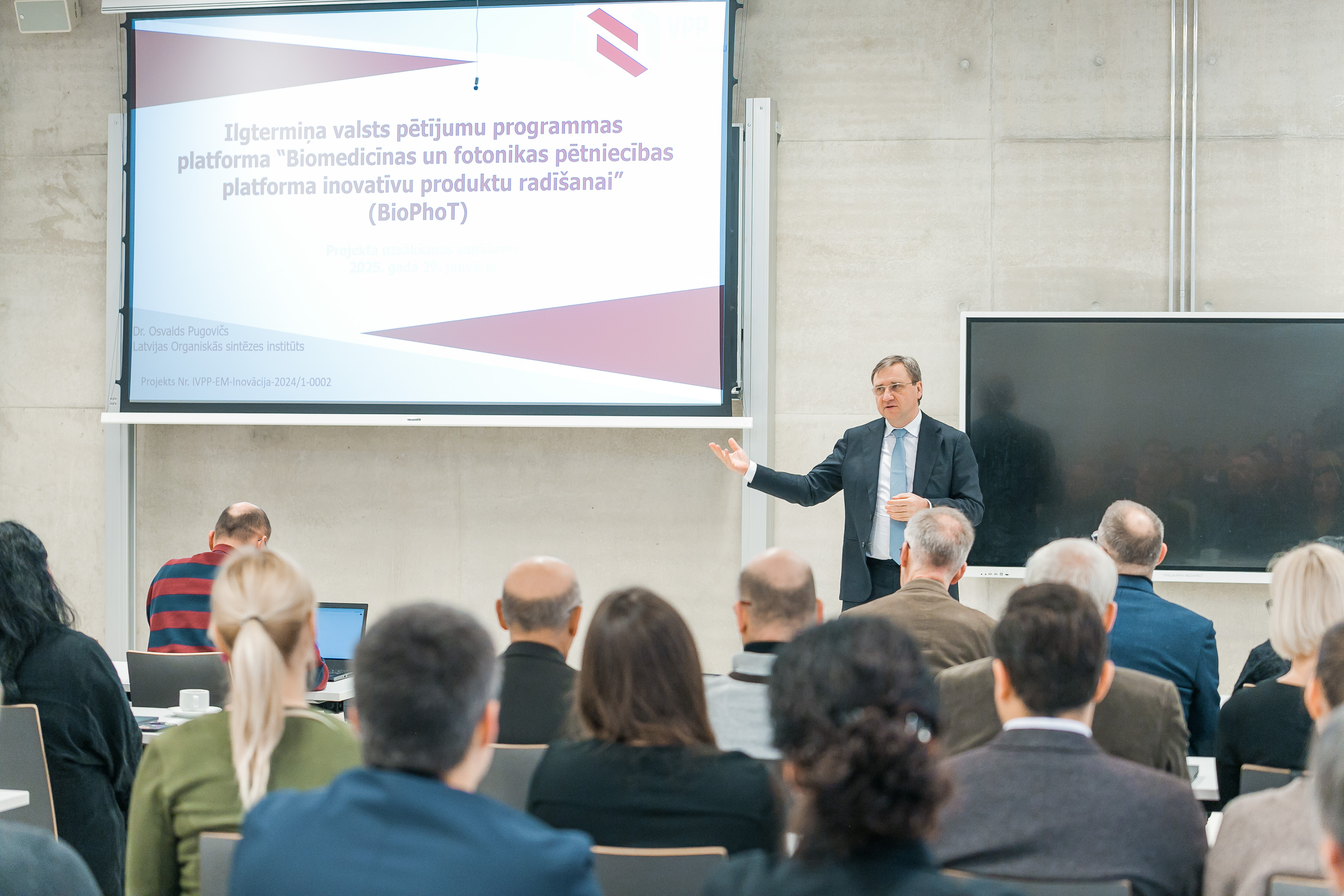 Cilvēki sapulcē, priekšpusē ekrāns un runātājs