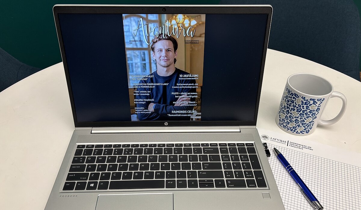 Uz galda novietots atvērts klēpjdators, kura ekrānā redzams žurnāla vāks ar nosaukumu “Avantūra”