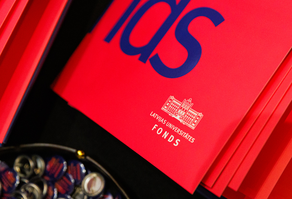 Sarkanas mapes ar Latvijas Universitātes Fonda logotipu un trauks ar nozīmītēm priekšplānā.