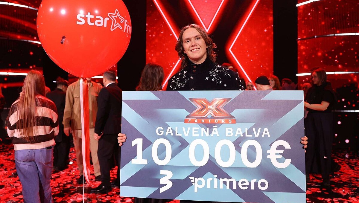 Uz skatuves redzams cilvēks, kas tur lielu čeku ar uzrakstu “X Faktors”, “Galvenā balva 10 000 €