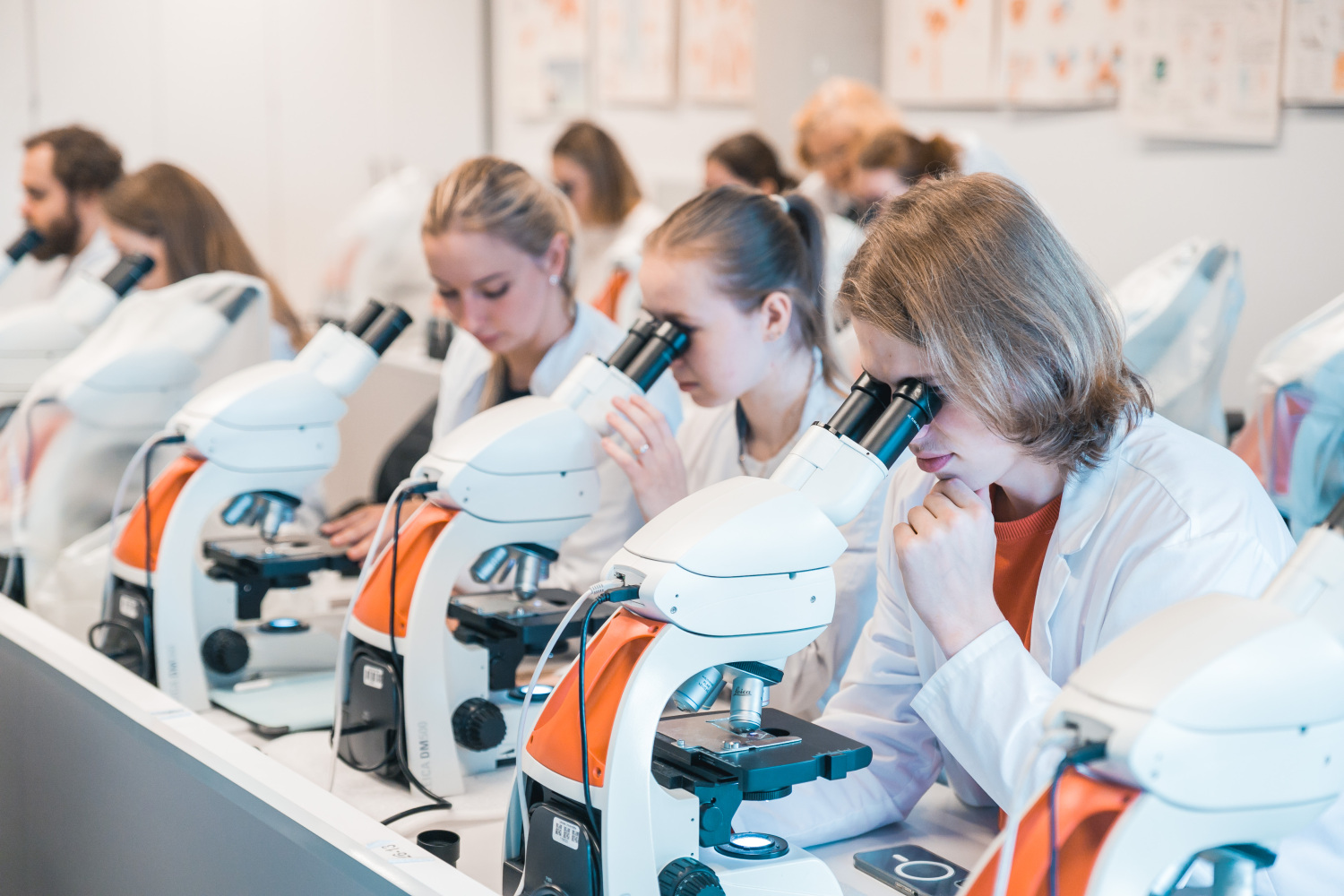 Cilvēki baltos halātos sēž pie galda un strādā ar mikroskopiem laboratorijas vidē. Fonā redzamas plakātu ilustrācijas uz sienas.