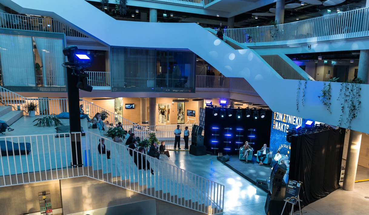 Centrā skatuve ar gaismu instalācijām un ekrānu, uz kura redzams teksts “Zinātnieku nakts”. Apkārt skatuvei sēž un stāv cilvēki, telpa izgaismota ar zilām un baltām gaismām.