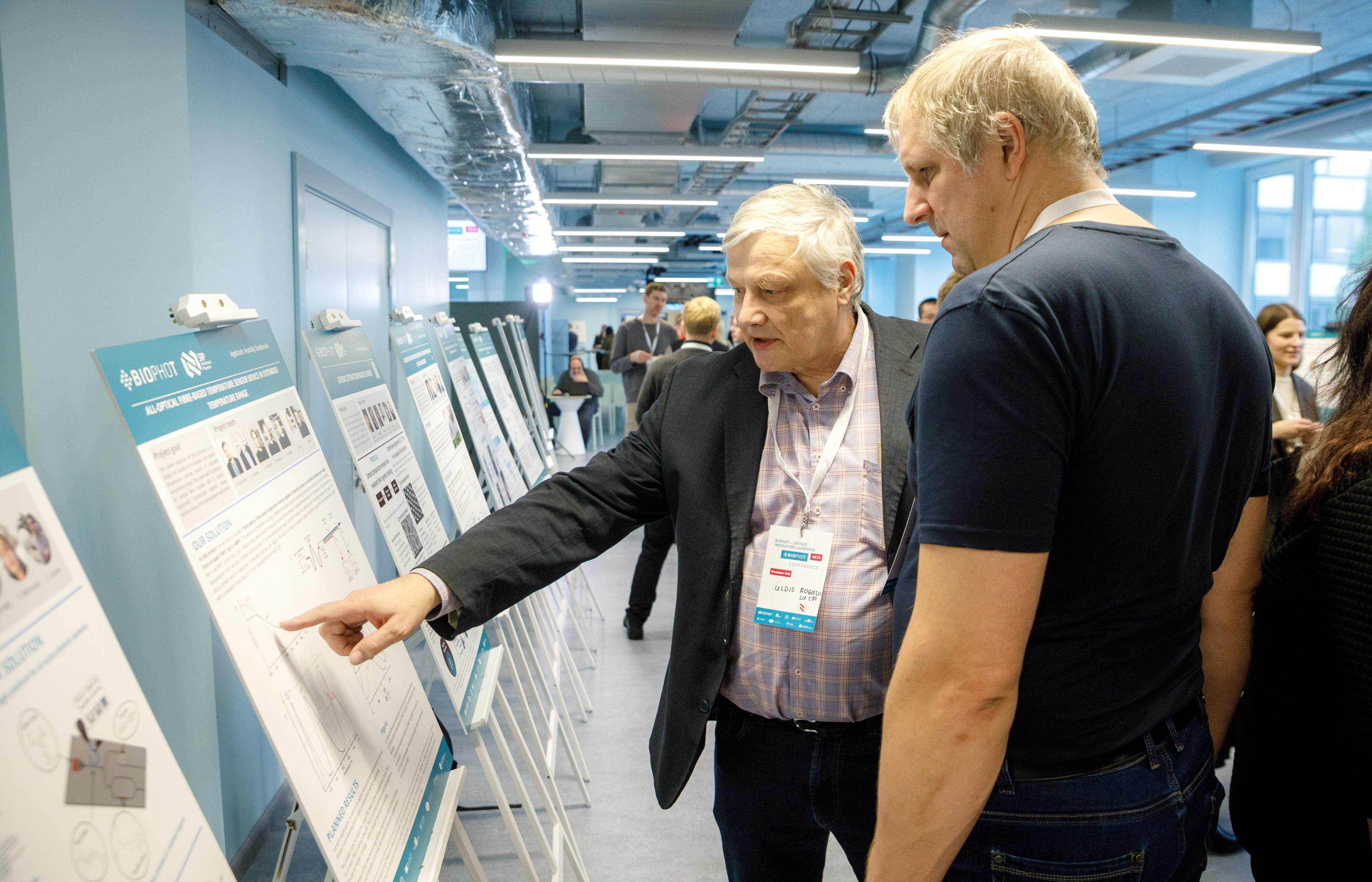 Zinātniskas konferences telpa, kur apmeklētāji aplūko un pārrunā pētījumu plakātus izstādē.