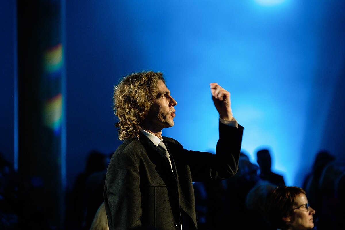 Persona stāv auditorijā un uzrunā klausītājus, paceļot roku; fonā redzama tumša telpa ar zilu skatuves apgaismojumu un klausītāju siluetiem.