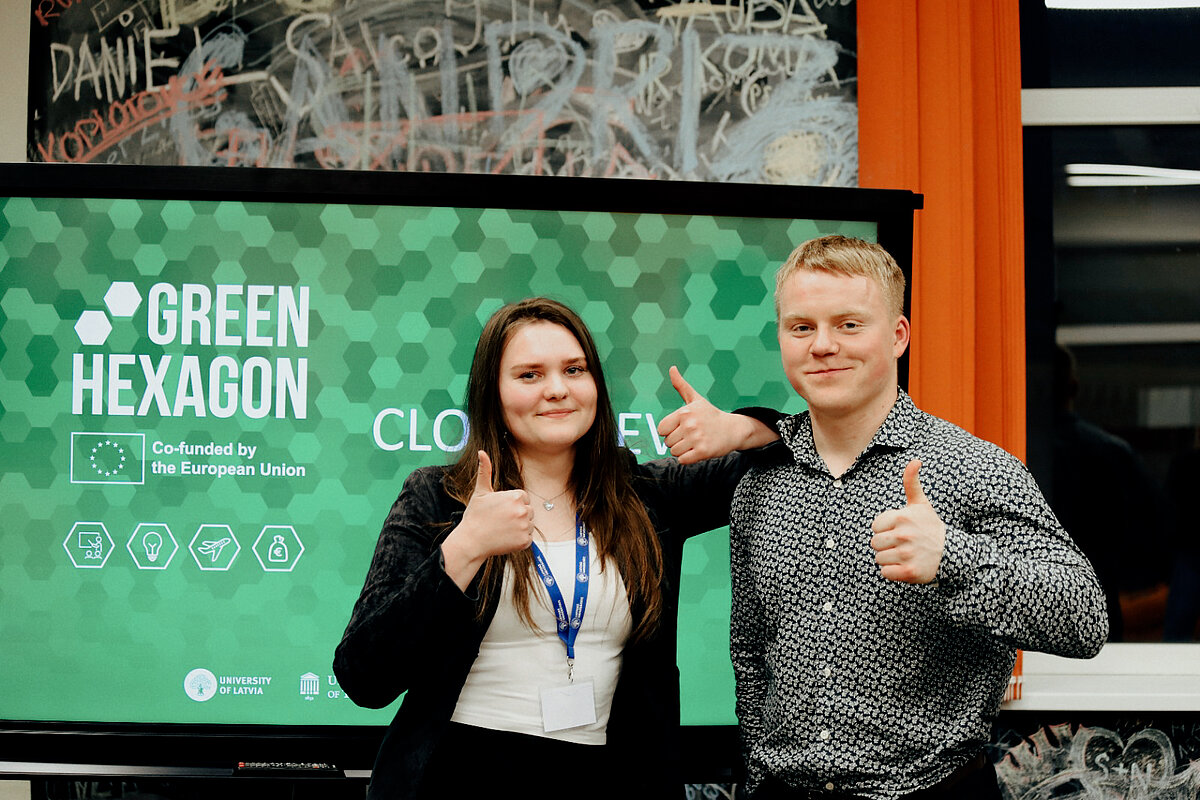 Divi cilvēki stāv pie GREEN HEXAGON ekrāna un rāda īkšķus uz augšu