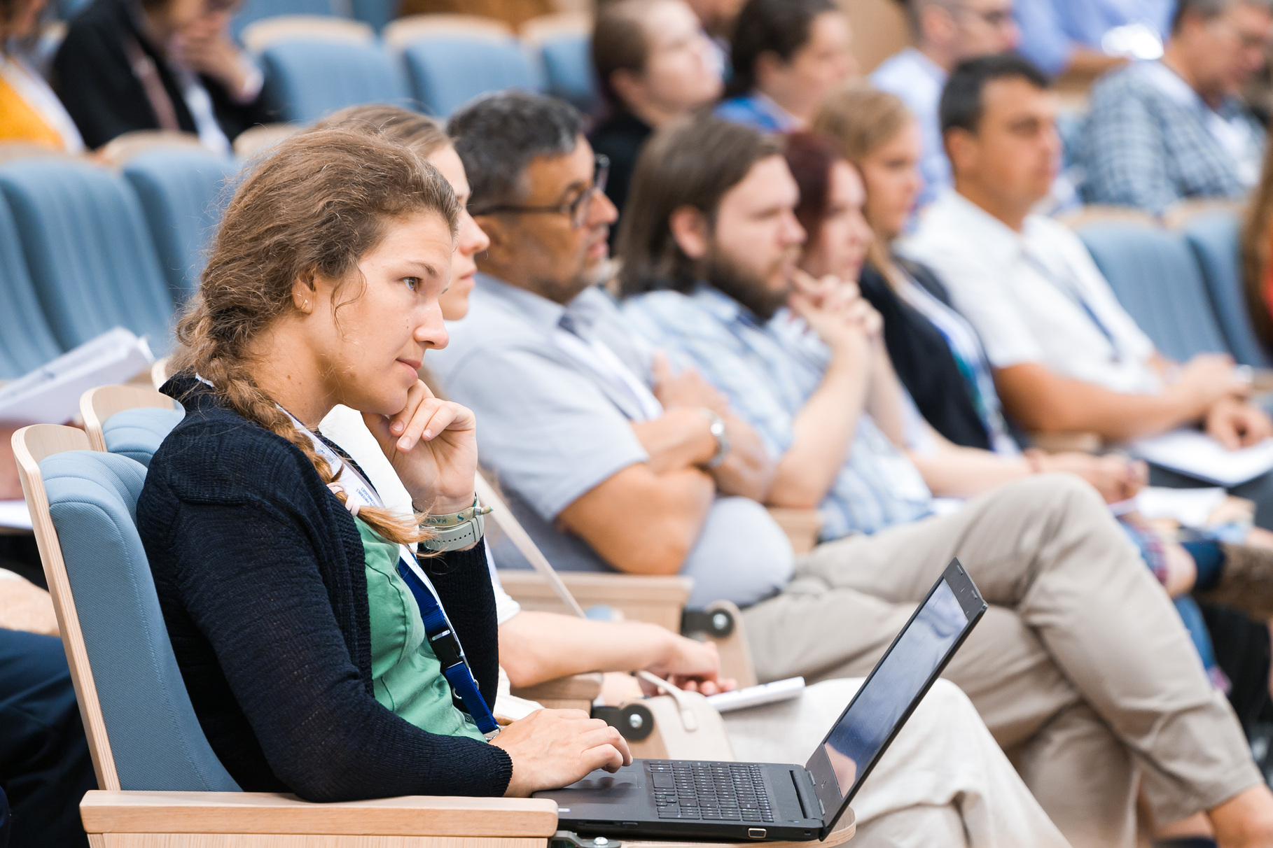 Konferenču zāles skatītāju rindās sēž dalībnieki, klausoties priekšlasījumā; priekšplānā persona ar klēpjdatoru uz ceļiem un piezīmju materiāliem blakus.
