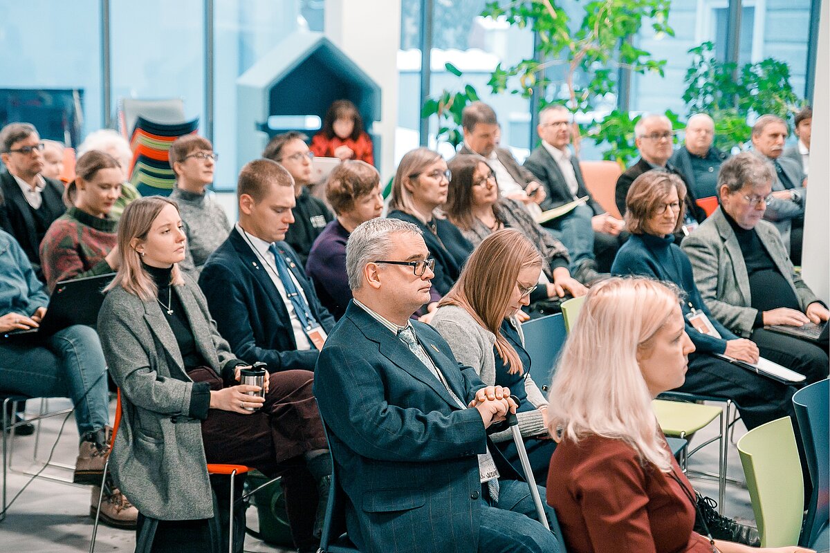 Semināra dalībnieki sēž rindās modernā iekštelpā un klausās prezentāciju; fonā redzamas telpaugi un gaišs interjers.
