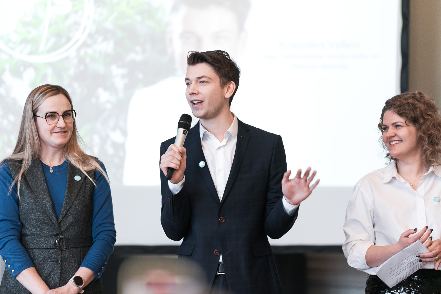 Jaunie skolotāji (3 cilvēki), vienam rokā mikrofons, fonā prezentācija