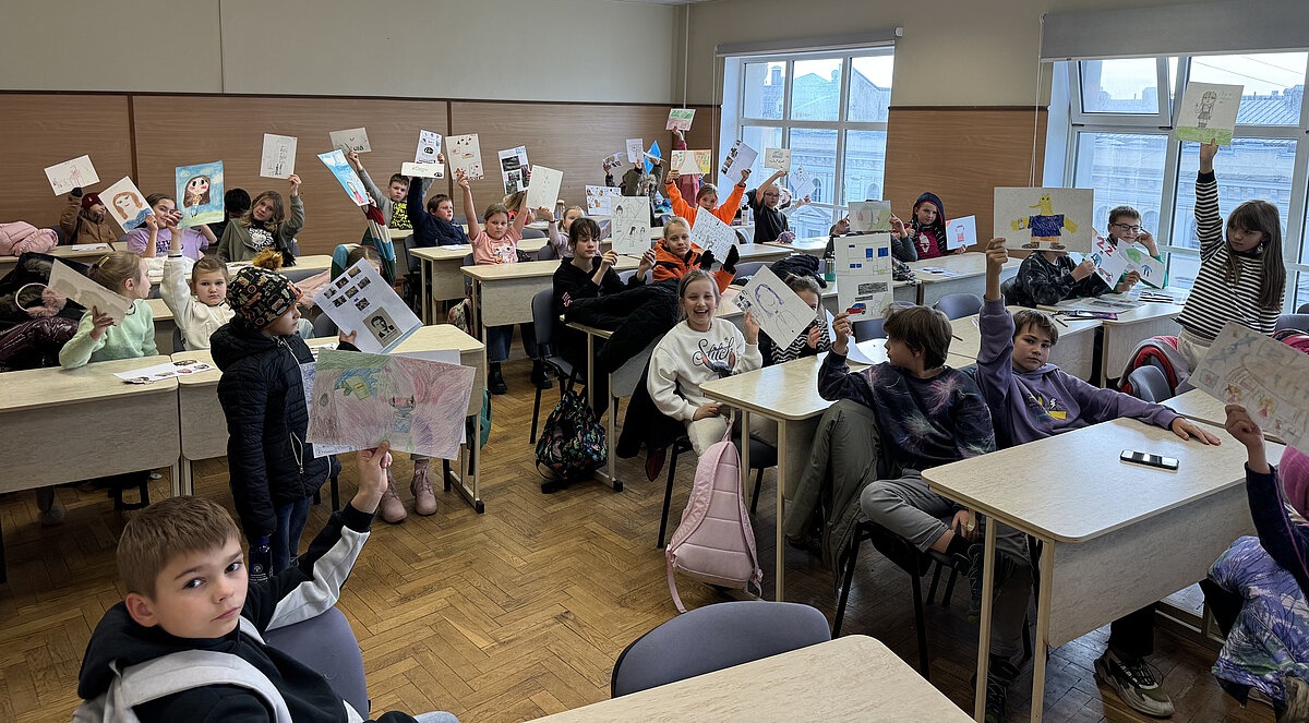 Klases telpā bērni sēž pie galdiem un ar paceltām rokām demonstrē savus zīmējumus un radošos darbus. Telpā redzami vairāki plakāti ar attēliem un ilustrācijām, pie logiem ieplūst dienasgaisma, radot gaišu un rosīgu atmosfēru