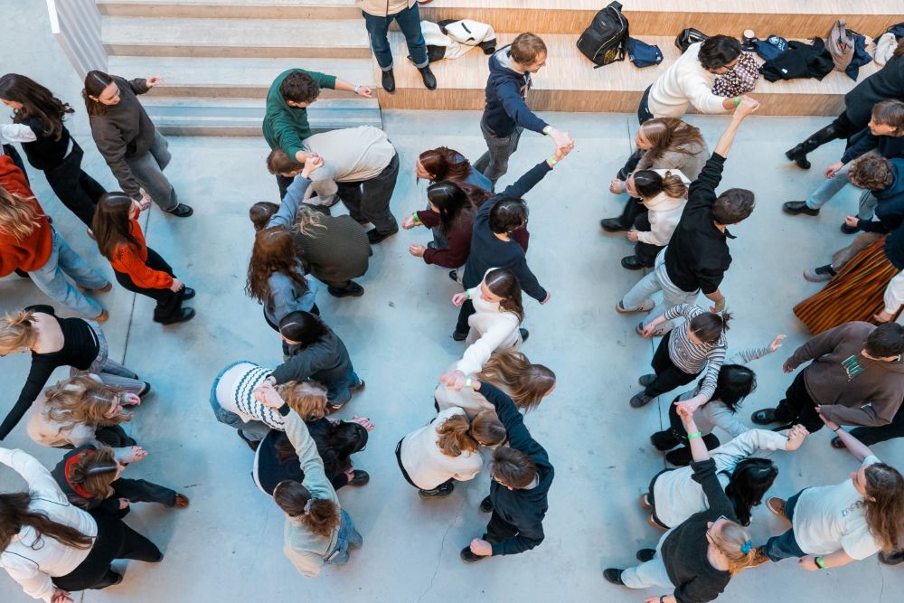 Skats no augšas uz lielu cilvēku grupu, kas telpā izkārtojušies vairākās rindās un savstarpēji satver rokas, veidojot cilpas un kustību rakstus. Apkārt redzamas kāpnes, somas un gaišas grīdas virsma
