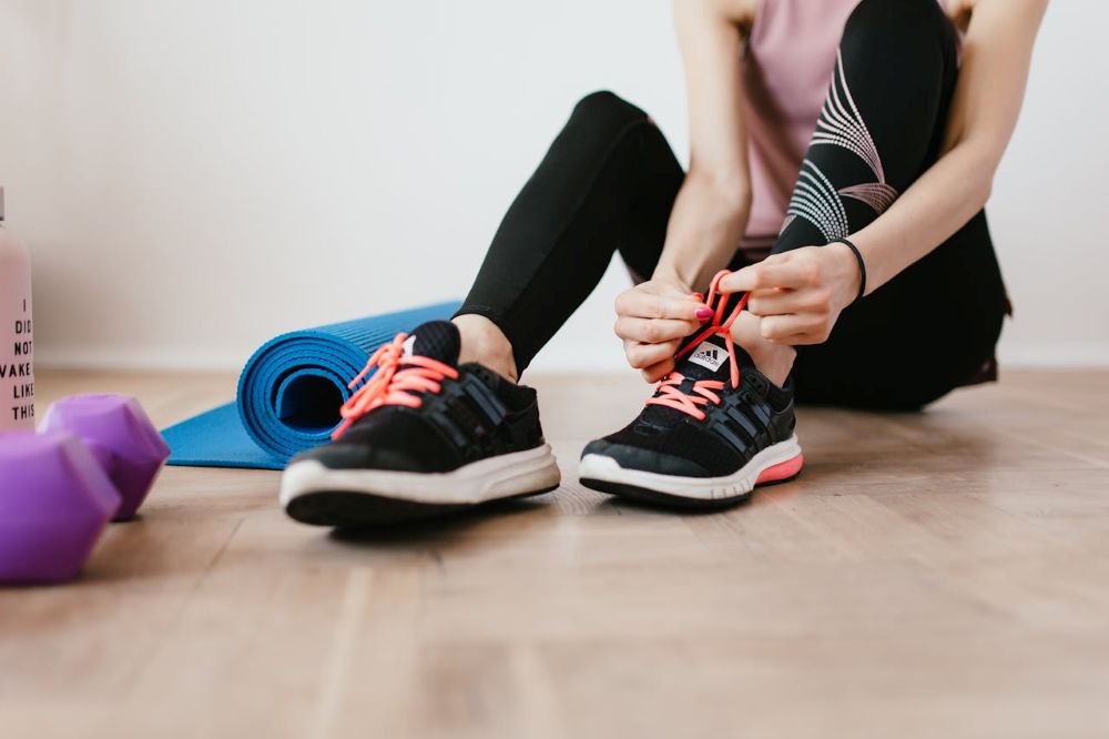 Cilvēks sporta apģērbā sēž uz grīdas un sasien sporta apavus. Blakus ir sarullēts vingrošanas paklājiņš un uz grīdas redzami rokas svari.