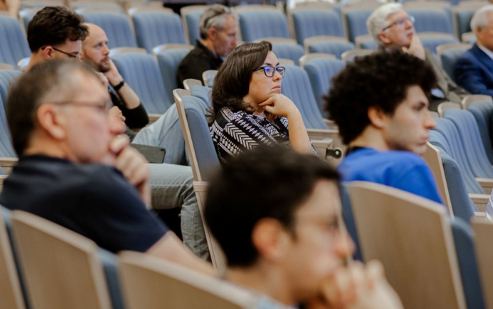Cilvēki sēž auditorijā rindās un klausās pasākumu vai prezentāciju. Redzamas vairākas krēslu rindas un zāles interjers ar gaišiem toņiem