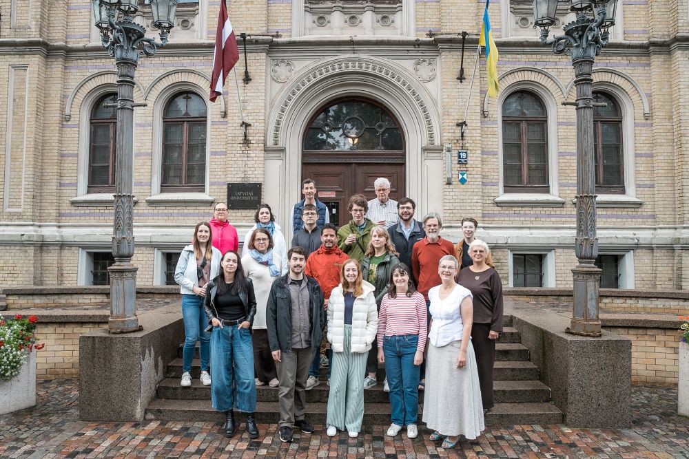 Grupas kopfoto pie LU ēkas fasādes ar dekoratīviem ķieģeļiem un arku ieeju. Cilvēki stāv vairākās rindās uz kāpnēm. Abās pusēs redzami dekoratīvi laternu stabi, un pie ēkas izkārtas Latvijas un Ukrainas karoga krāsas