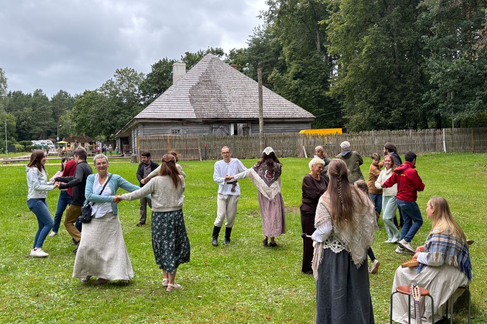 Cilvēku grupa brīvdabas vidē veido loku un dejo tradicionālu rotaļdeju zaļā laukumā. Daļa dalībnieku tērpušies vēsturiskos vai etnogrāfiski iedvesmotos tērpos. Fonā redzama koka ēka ar stāvu jumtu un koka žogs