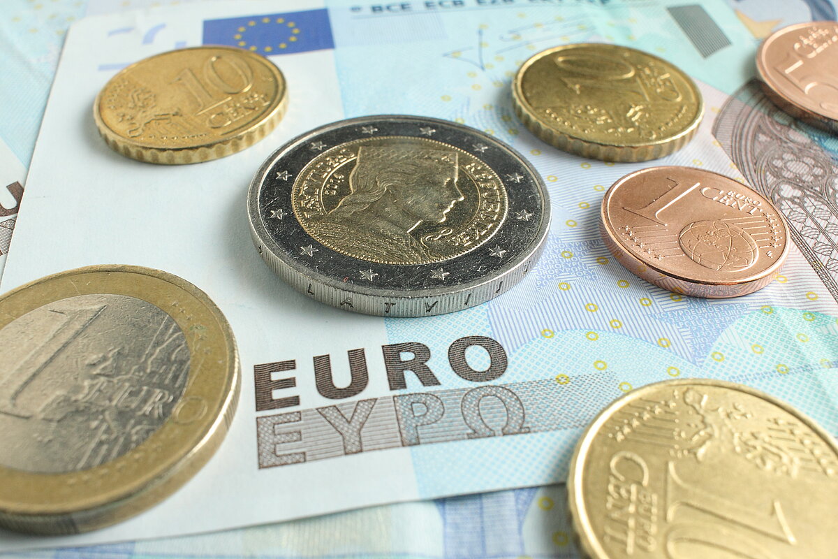 Eiro banknote ar uzrakstu “EURO” un vairākām eiro monētām dažādos nominālos, kas izkārtotas uz banknotes virsmas tuvplānā.