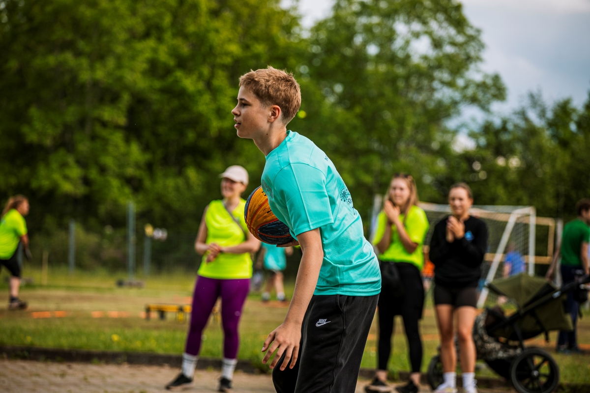 Āra sporta aktivitāte: bērns stāv priekšplānā ar bumbu rokās, gatavojoties darbībai, fonā redzami citi dalībnieki un treniņa zona ar sporta inventāru zaļā parkā.