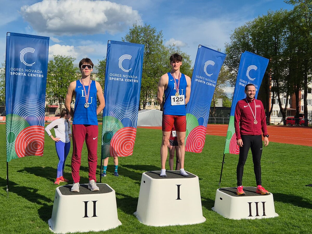 Trīs sportisti stāv uz uzvarētāju pjedestāla stadiona zālienā, valkā medaļas un sporta formu; centrā pirmajā vietā (I), pa kreisi otrā vieta (II), pa labi trešā vieta (III). Fonā redzami “Ogres novada sporta centrs” karogi, skrejceļš un koki.