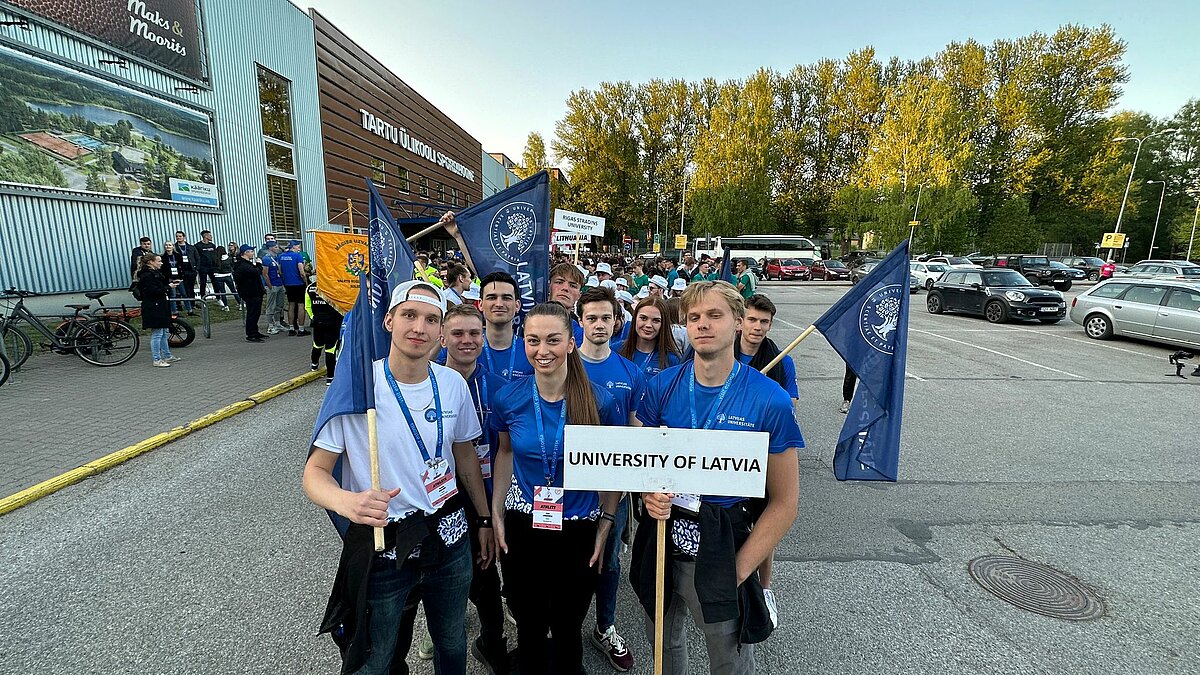 Latvijas Universitātes studentu komanda zilos krekliņos piedalās SELL studentu sporta spēļu gājienā, turot karogus un zīmi “University of Latvia”; fonā redzama pasākuma norises vieta pie ēkas ar uzrakstu “Tartu Ülikool”, citi dalībnieki un automašīnas.