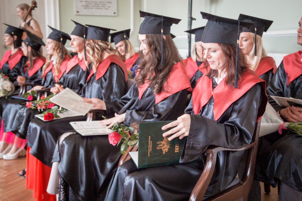  Absolventi akadēmiskajos tērpos sēž izlaiduma ceremonijā, rokās turot diplomus un ziedus.