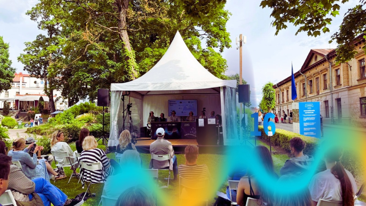 Brīvdabas diskusija festivālā “LAMPA”, kur klausītāji sēž krēslos zem kokiem un vēro sarunu uz skatuves zem baltas nojumes. Pasākums notiek pilsētvidē, apkārt redzami koki, ēkas un informatīvas norādes.
