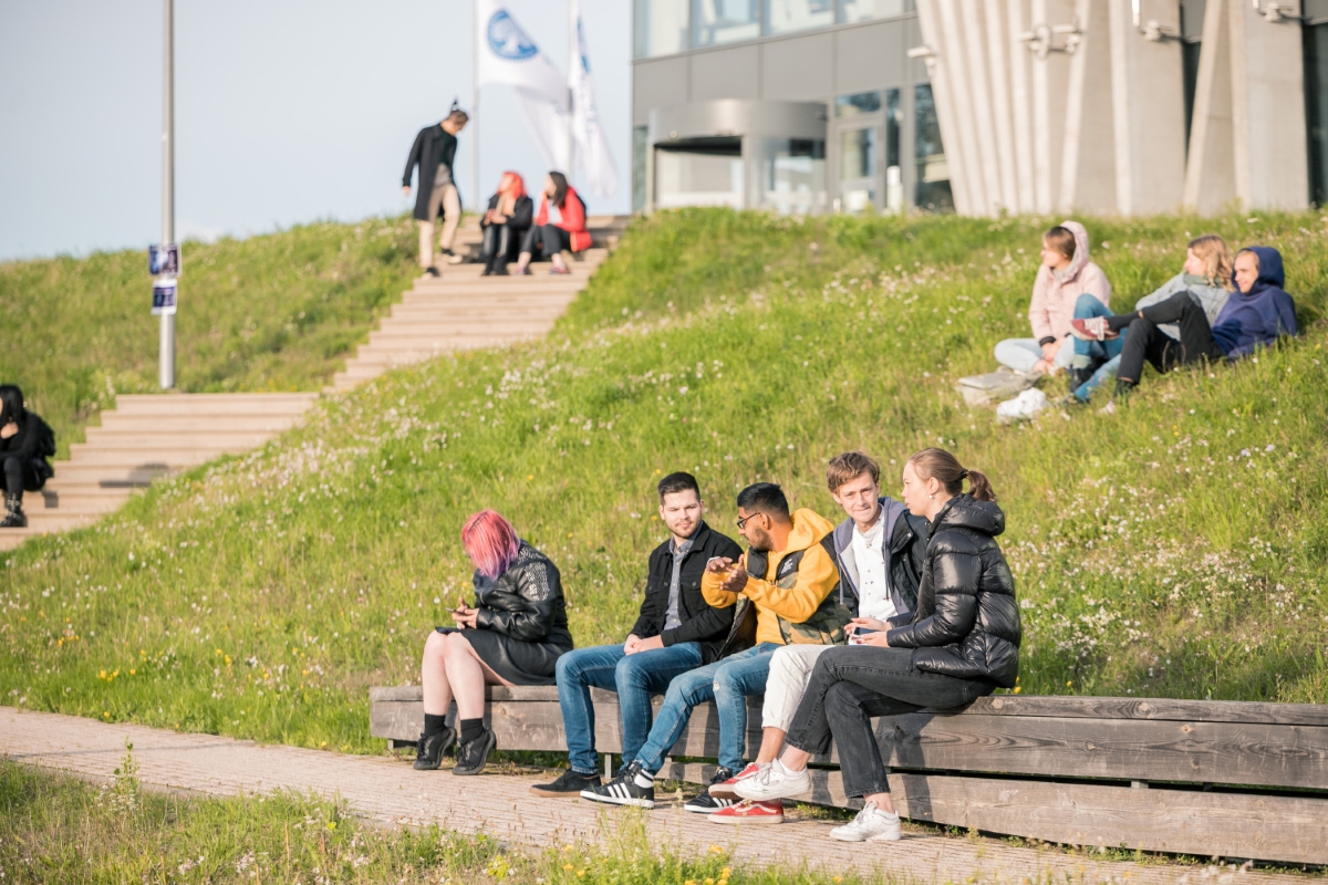 Cilvēki sēž uz soliņiem un zālienā pie Latvijas Universitātes ēkas saulainā dienā.