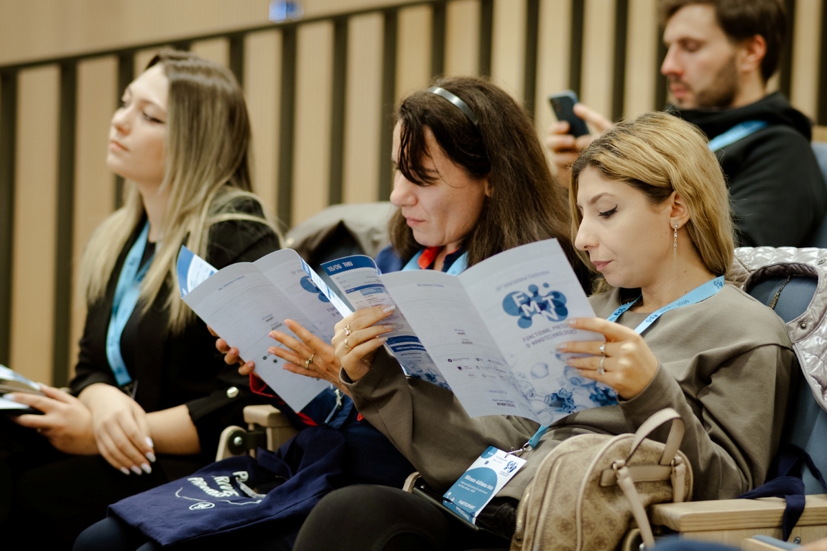 Pasākuma apmeklētāji sēž auditorijā un lasa konferences programmas bukletus