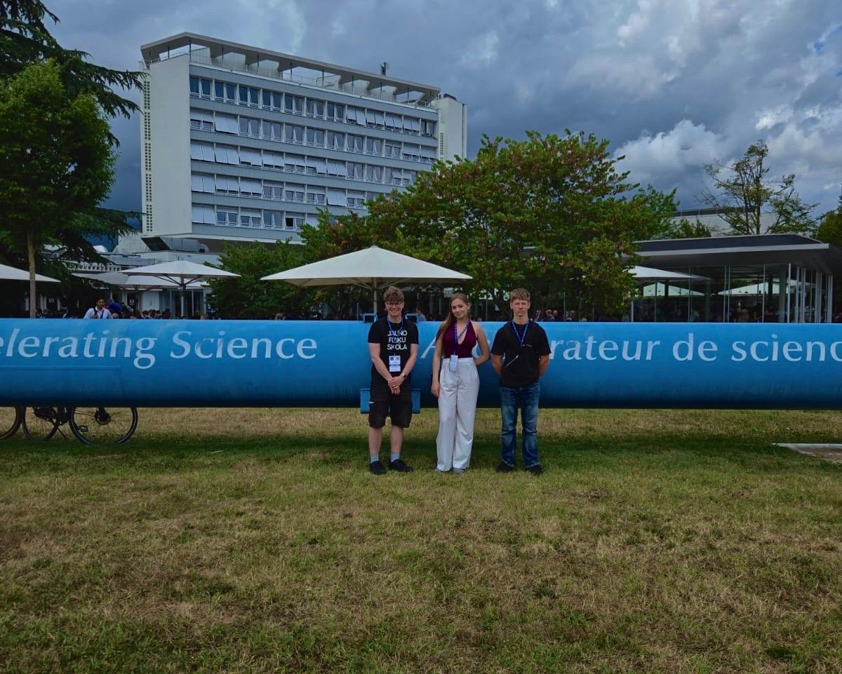 **ALT teksts:**  Trīs jaunieši ar dalībnieku kartēm stāv zālienā pie lielas zilas instalācijas ar uzrakstu “Accelerating Science”. Fonā redzama moderna ēka, koki un pasākuma teritorija zem mākoņainām debesīm.