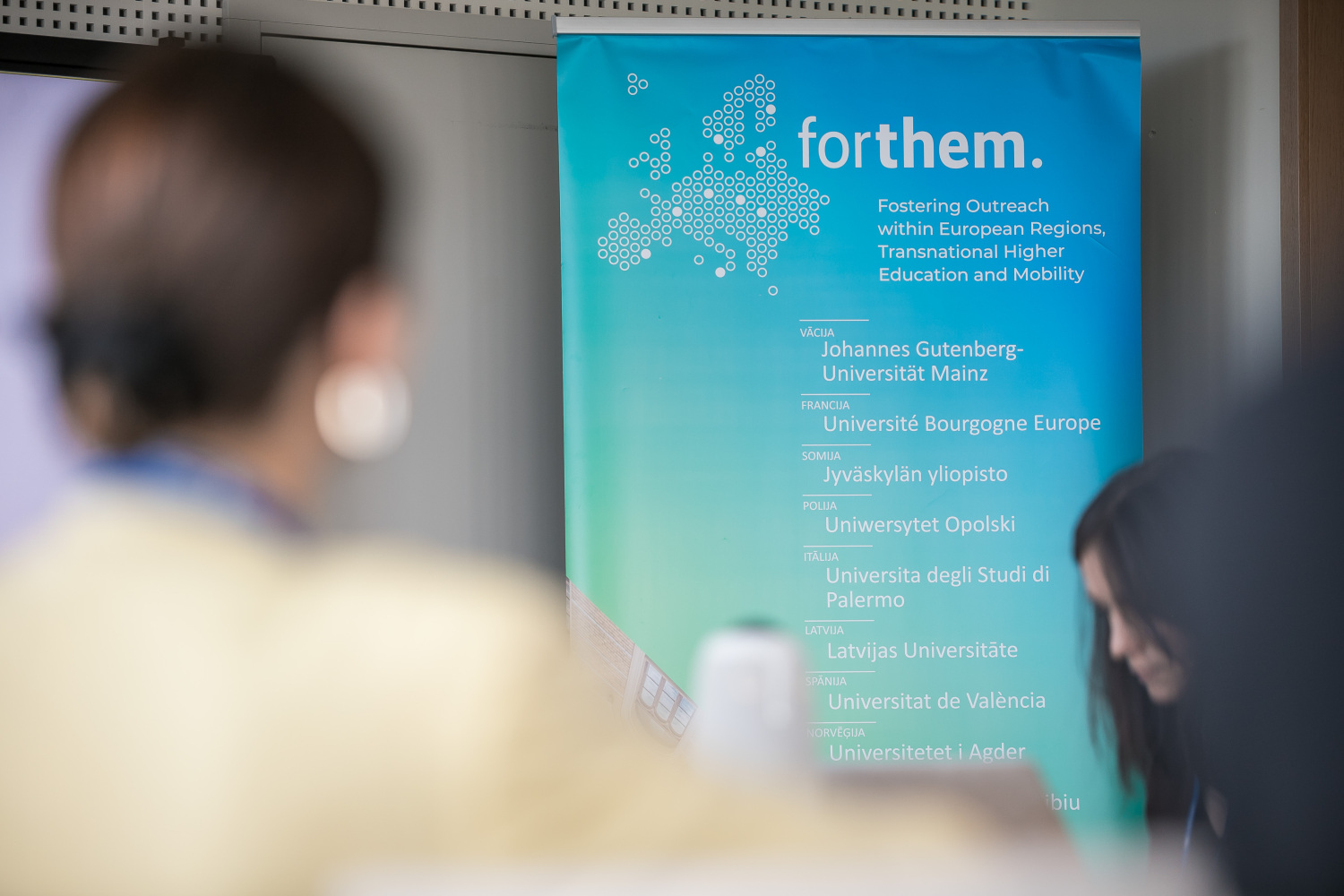 Konferenču telpā redzams “FORTHEM” projekta stends ar Eiropas kartes grafiku un partneruniversitāšu nosaukumiem. Priekšplānā un fonā redzami pasākuma dalībnieki ārpus fokusa.