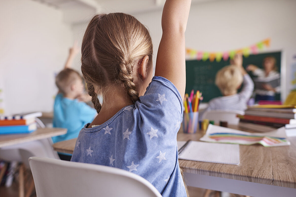 Meitenīte ar paceltu roku klasē. Fonā tāfele. 