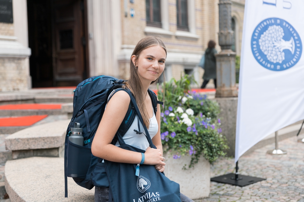 Persona ar mugursomu un Latvijas Universitātes somu sēž pie universitātes galvenās ēkas ieejas. Blakus redzams Latvijas Universitātes karogs un ziedu kompozīcija.