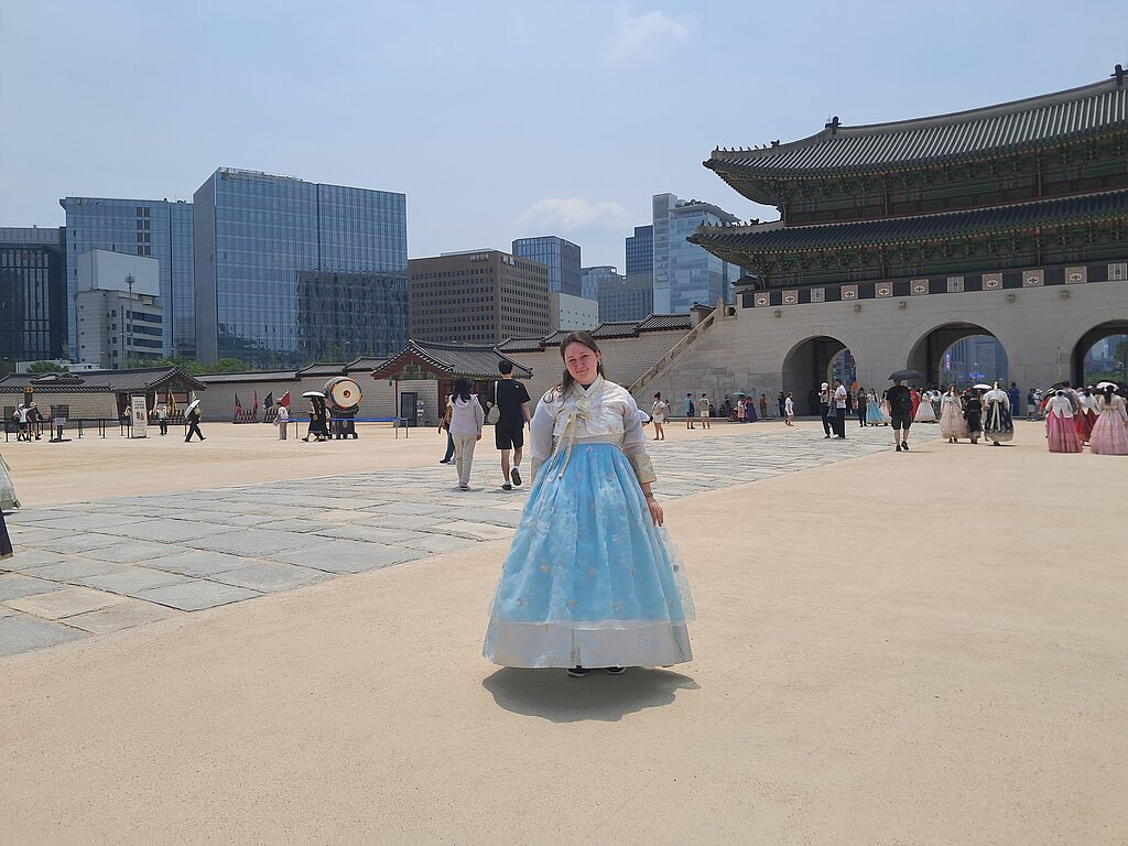 **ALT teksts:**  Persona tradicionālā korejiešu hanbok tērpā stāv plašā laukumā pie vēsturiskas pils kompleksa. Fonā redzama grezni rotāta pils ēka, apmeklētāji un mūsdienu augstceltnes, kas kontrastē ar vēsturisko arhitektūru.