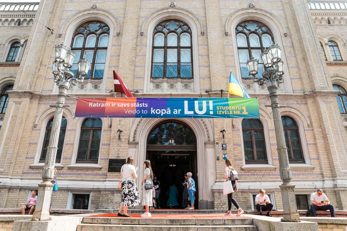 Cilvēki pie Latvijas Universitātes galvenās ēkas ieejas, virs kuras izvietots reklāmas baneris par studijām LU.