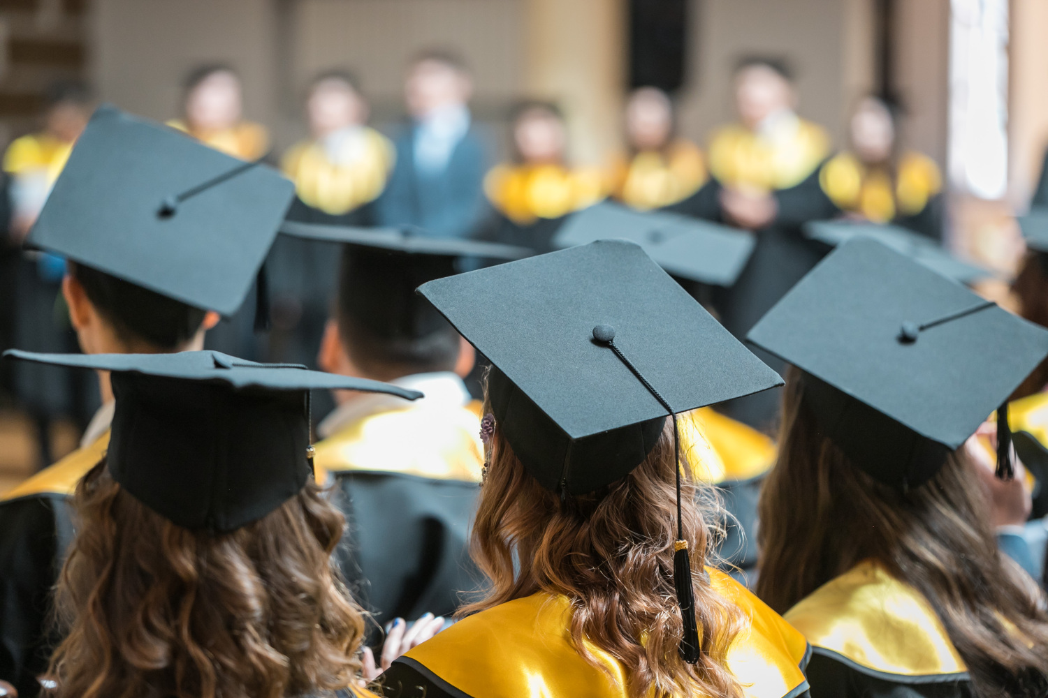 Studentu grupa ar akadēmiskajām cepurēm un talāriem sēž ceremonijas laikā, vērsti pret skatuvi, kur norisinās izlaiduma pasākums