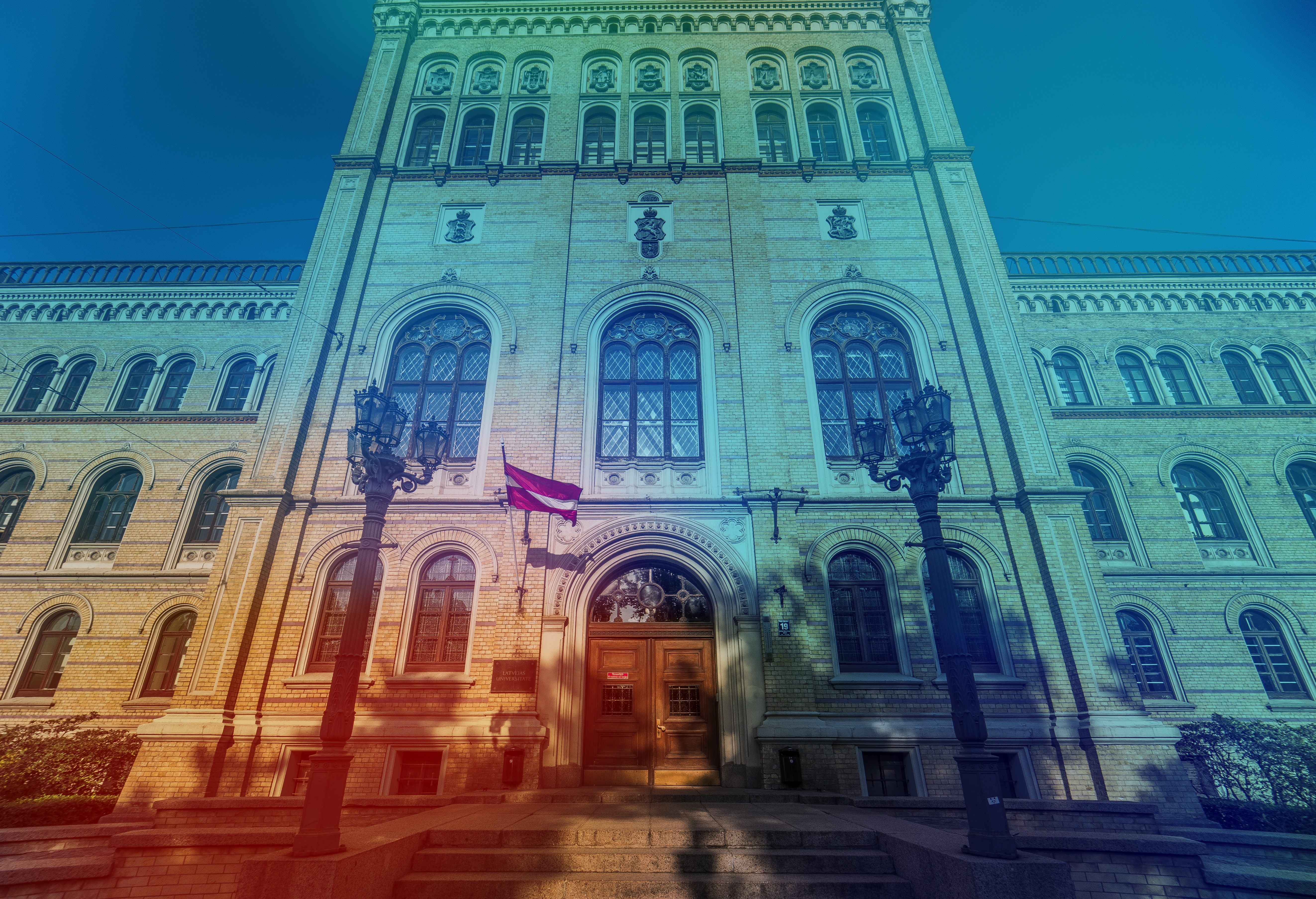 Latvijas Universitātes galvenās ēkas fasāde Rīgā ar Latvijas karogu pie ieejas. Vēsturiskā ķieģeļu ēka ar dekoratīviem arhitektūras elementiem un arku logiem, apstrādāta ar zilzaļu krāsu filtru.