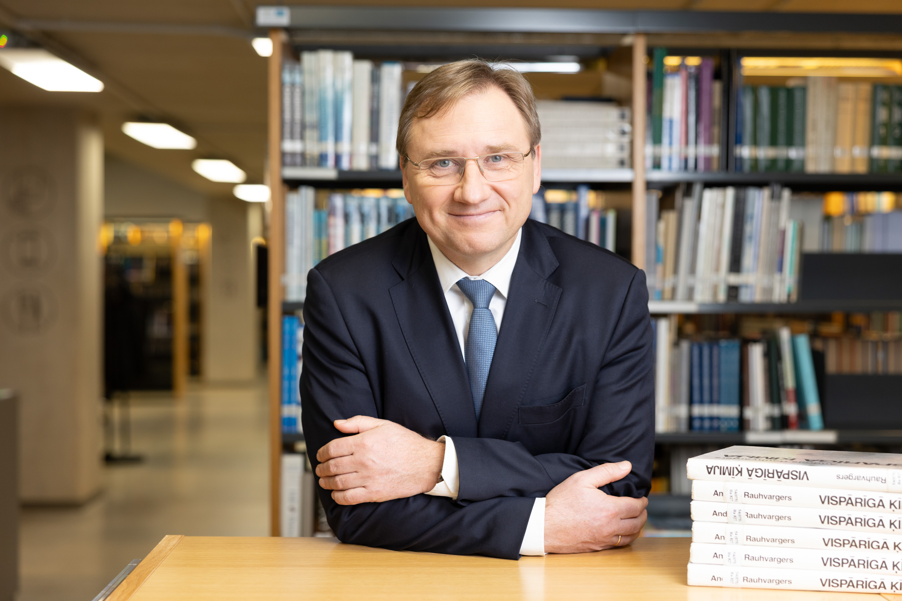LU rektors Gundars Bērziņš ar brillēm tumšā uzvalkā un zilā kaklasaitē sēž pie galda bibliotēkā. Blakus novietotas grāmatas ar uzrakstu "Vispārīgā ķīmija". Fonā grāmatplaukti ar zinātnisko literatūru.