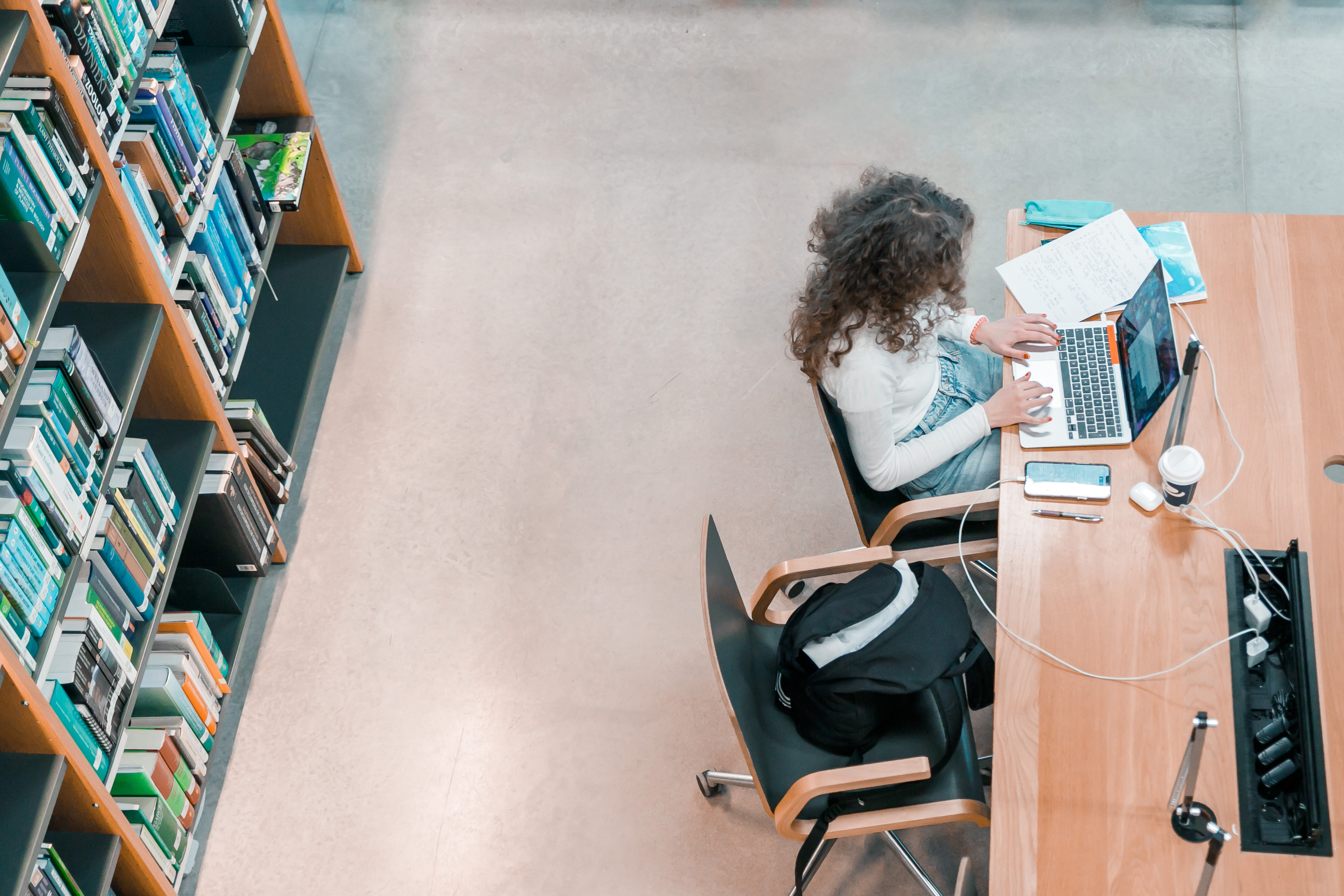 Studente sēž Latvijas Universitātes bibliotēkā pie galda, strādā pie portatīvā datora, uz galda ir piezīmju klade