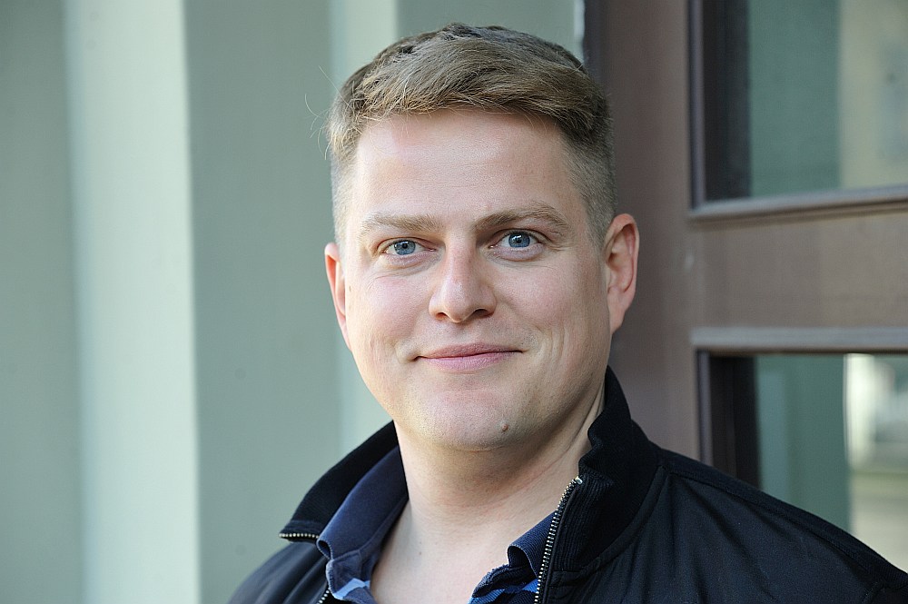 Oskars Gruziņš portretfoto melnā jakā, gaišā ārtelpu fonā.