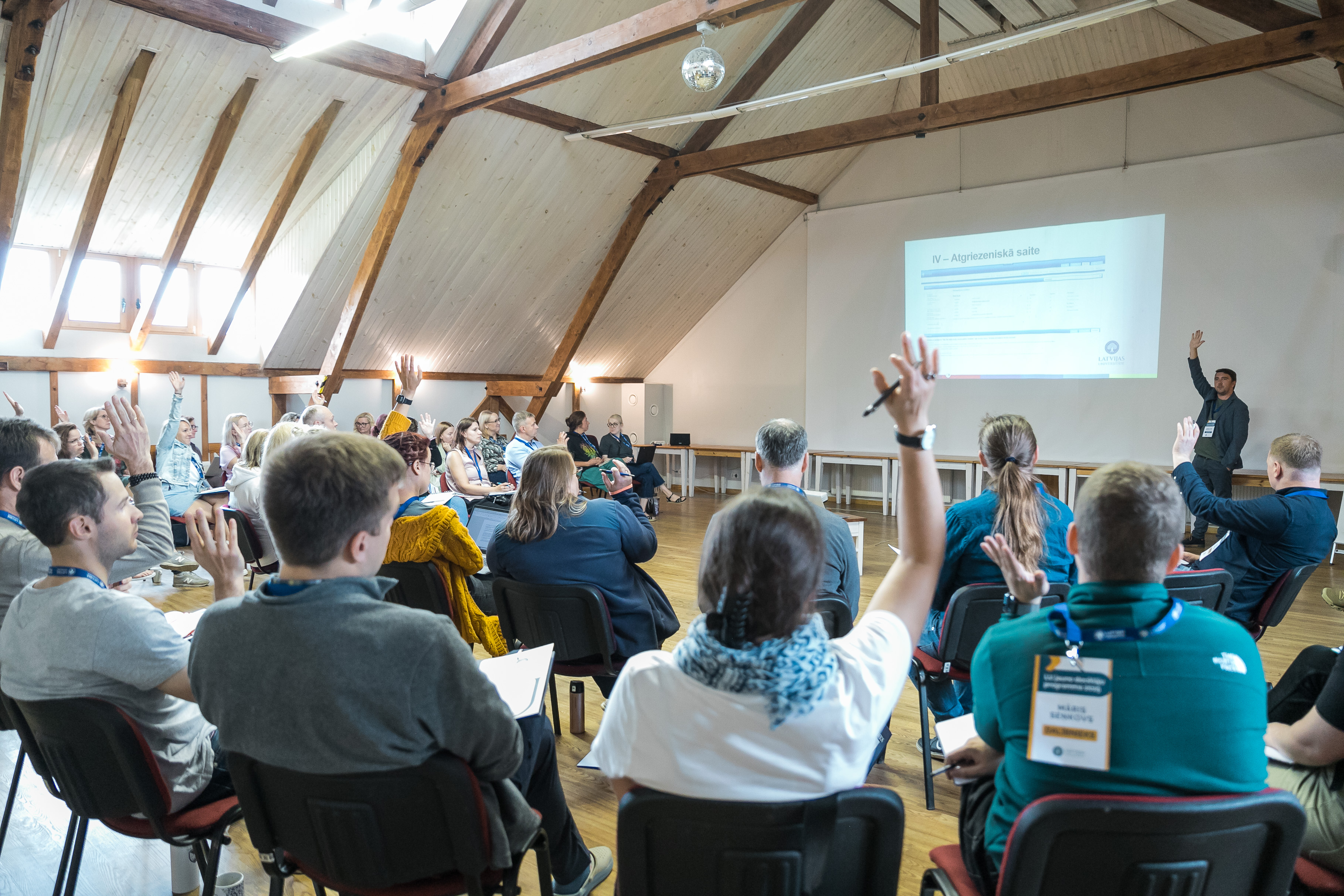Seminar or training participants raising their hands, seated on chairs in a bright room with wooden beams. A presenter stands by a screen displaying a slide titled "Atgriezeniskā saite" (Feedback). Participants are wearing name badge lanyards.