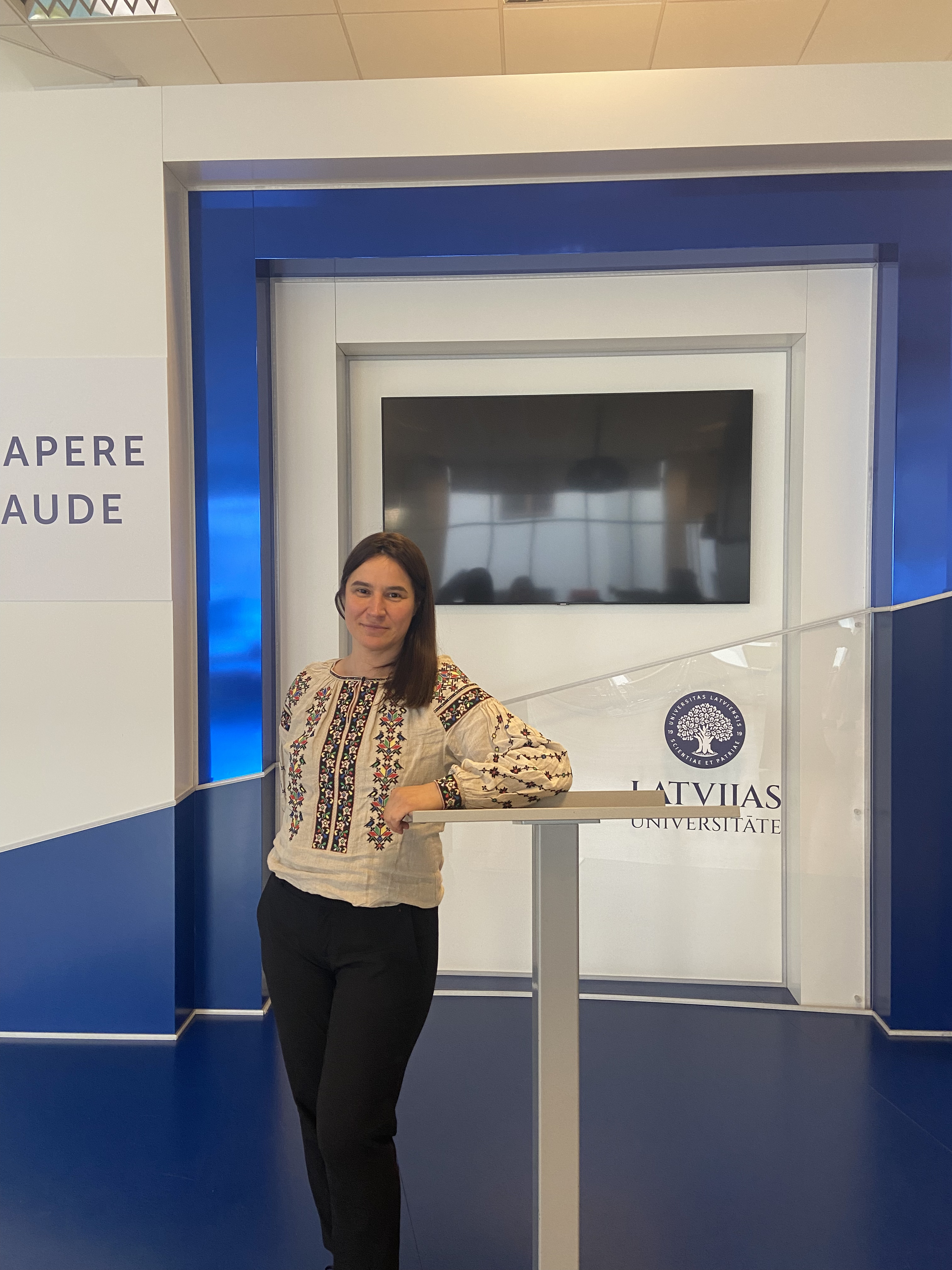 Hanna Markova baltā izšūtā blūzē stāv pie Latvijas Universitātes stenda ar universitātes logotipu un uzrakstu "Sapere aude". Fonā ekrāns un zilbalta dizaina fons.