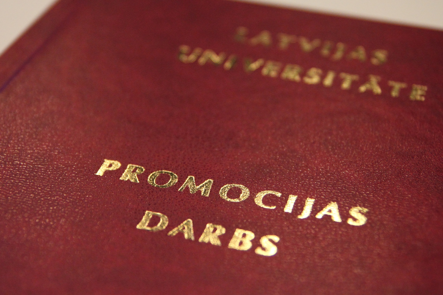 Latvijas Universitātes promocijas darbs ar tumši sarkanu vāku un zeltītiem uzrakstiem "Latvijas Universitāte. Promocijas darbs", novietots starp atvērtām grāmatām.