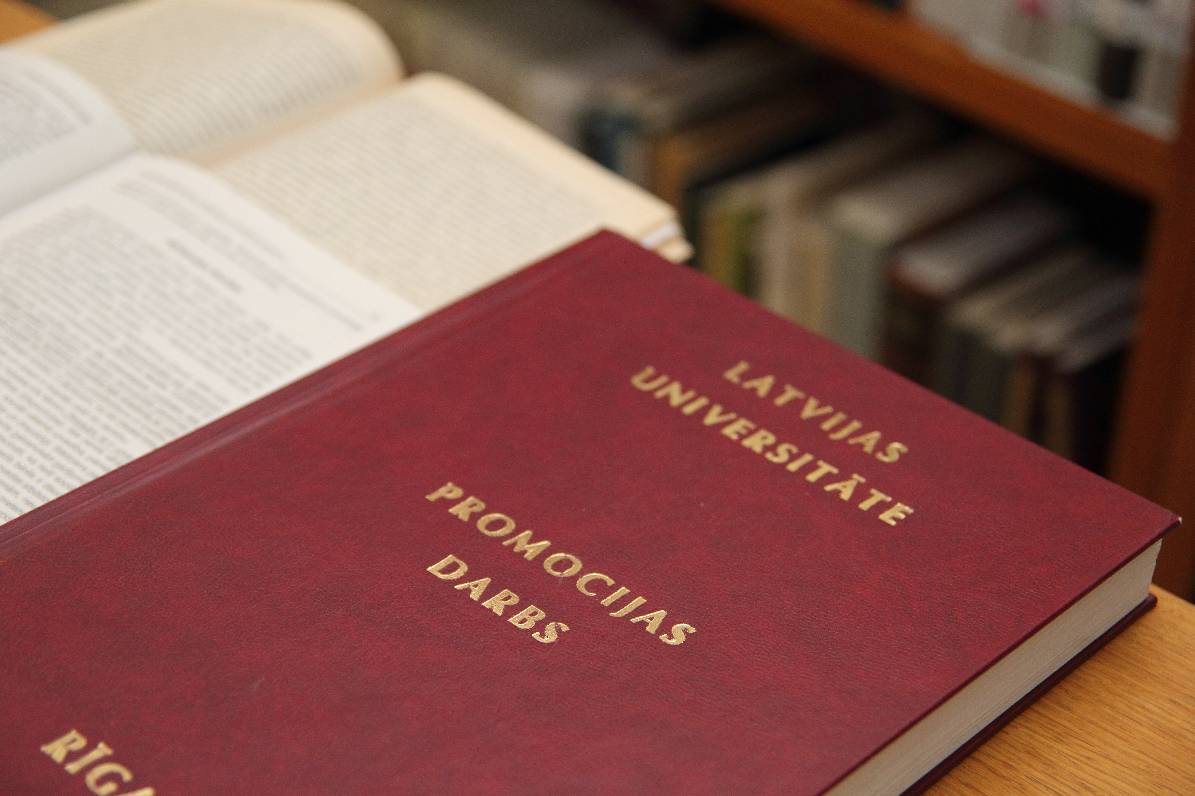 Latvijas Universitātes promocijas darbs ar tumši sarkanu vāku un zeltītiem uzrakstiem "Latvijas Universitāte. Promocijas darbs", novietots starp atvērtām grāmatām.