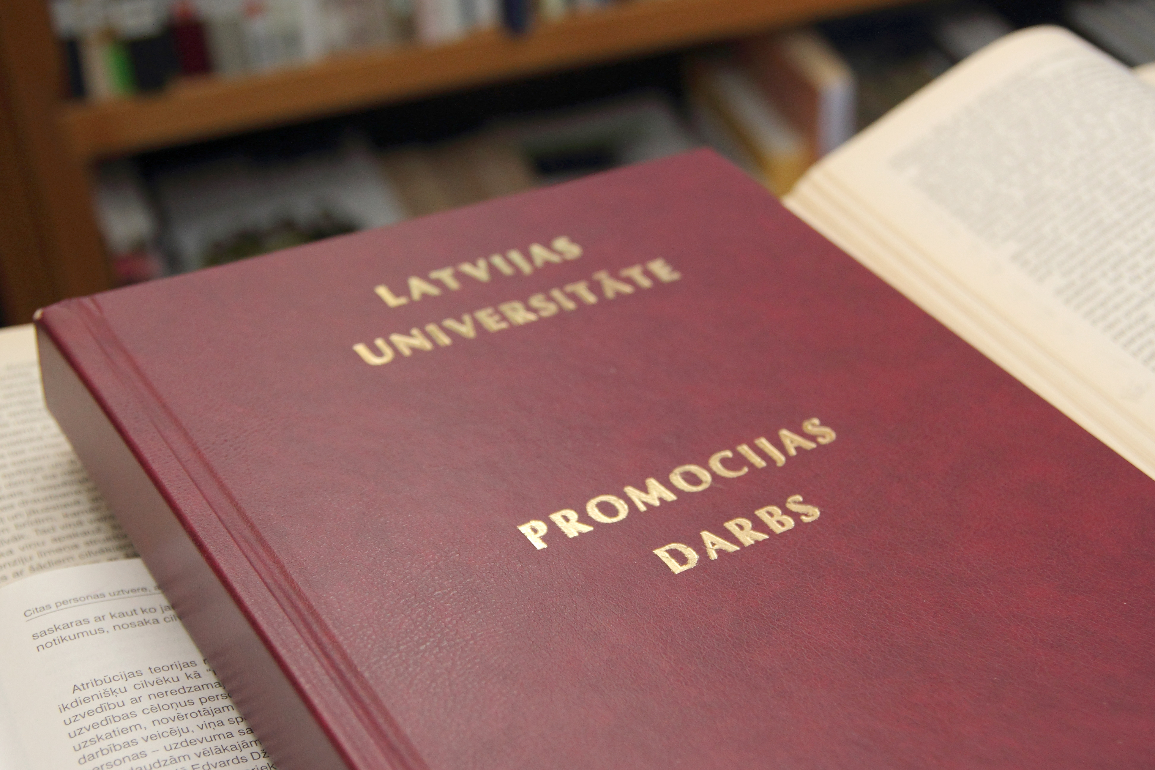 Latvijas Universitātes promocijas darbs ar tumši sarkanu vāku un zeltītiem uzrakstiem "Latvijas Universitāte. Promocijas darbs", novietots starp atvērtām grāmatām.