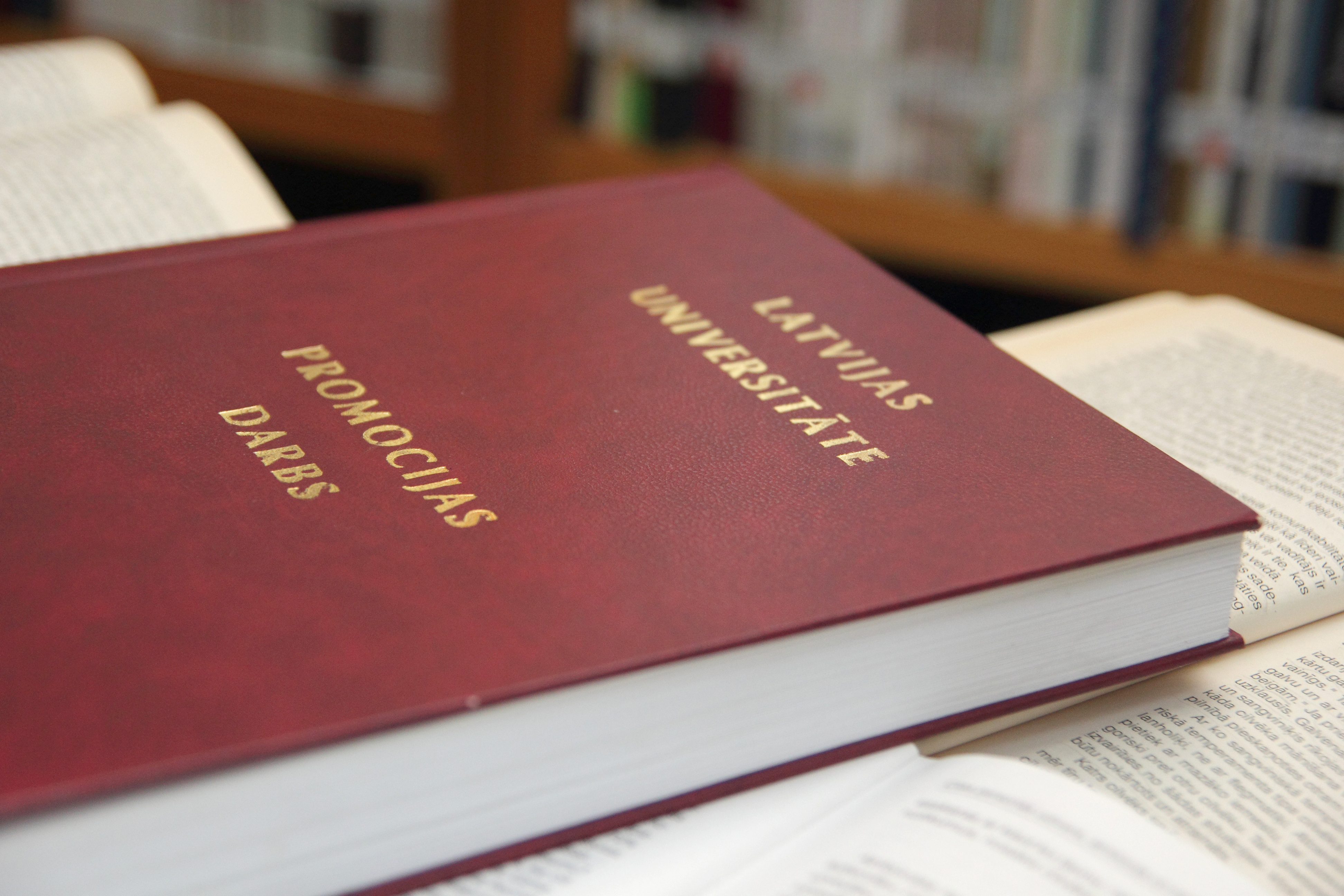 Latvijas Universitātes promocijas darbs ar tumši sarkanu vāku un zeltītiem uzrakstiem "Latvijas Universitāte. Promocijas darbs", novietots starp atvērtām grāmatām.
