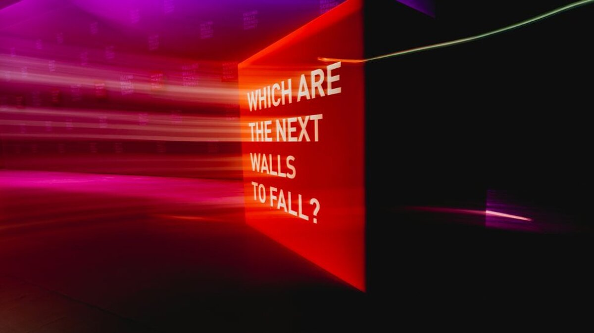 n a dark room, a brightly illuminated red wall is visible, featuring the English inscription “Which are the next walls to fall?”. Surrounding it are motion-created light trails in shades of pink and violet, creating a dynamic, futuristic atmosphere