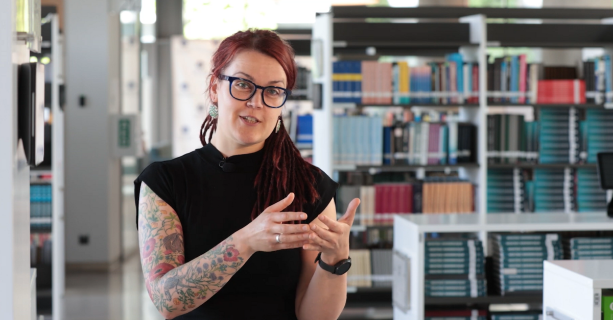 Vegas Polinska ar brillēm, sarkanbrūniem drediem un tetovējumiem uz rokas runā un žestikulē bibliotēkā. Fonā redzami grāmatplaukti ar krāsainām grāmatām.