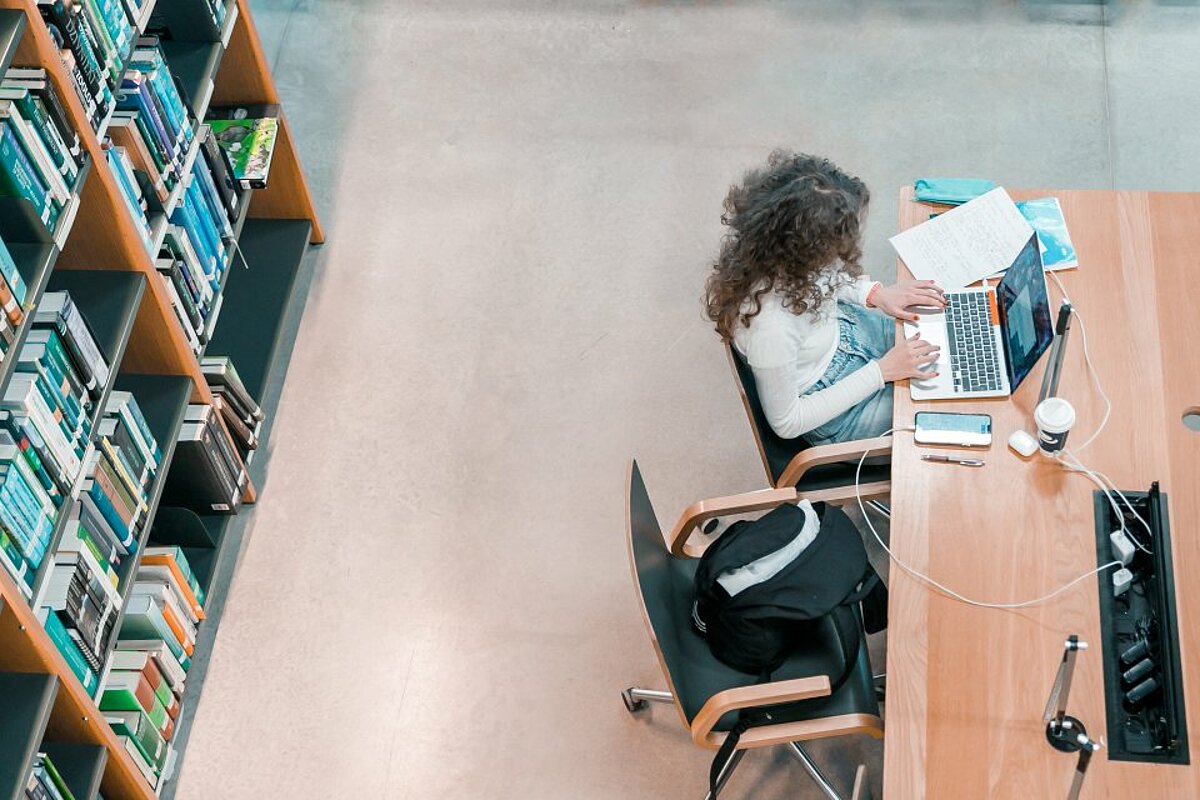 Overhead view of a student working at a computer in a library, with colourful bookshelves alongside.