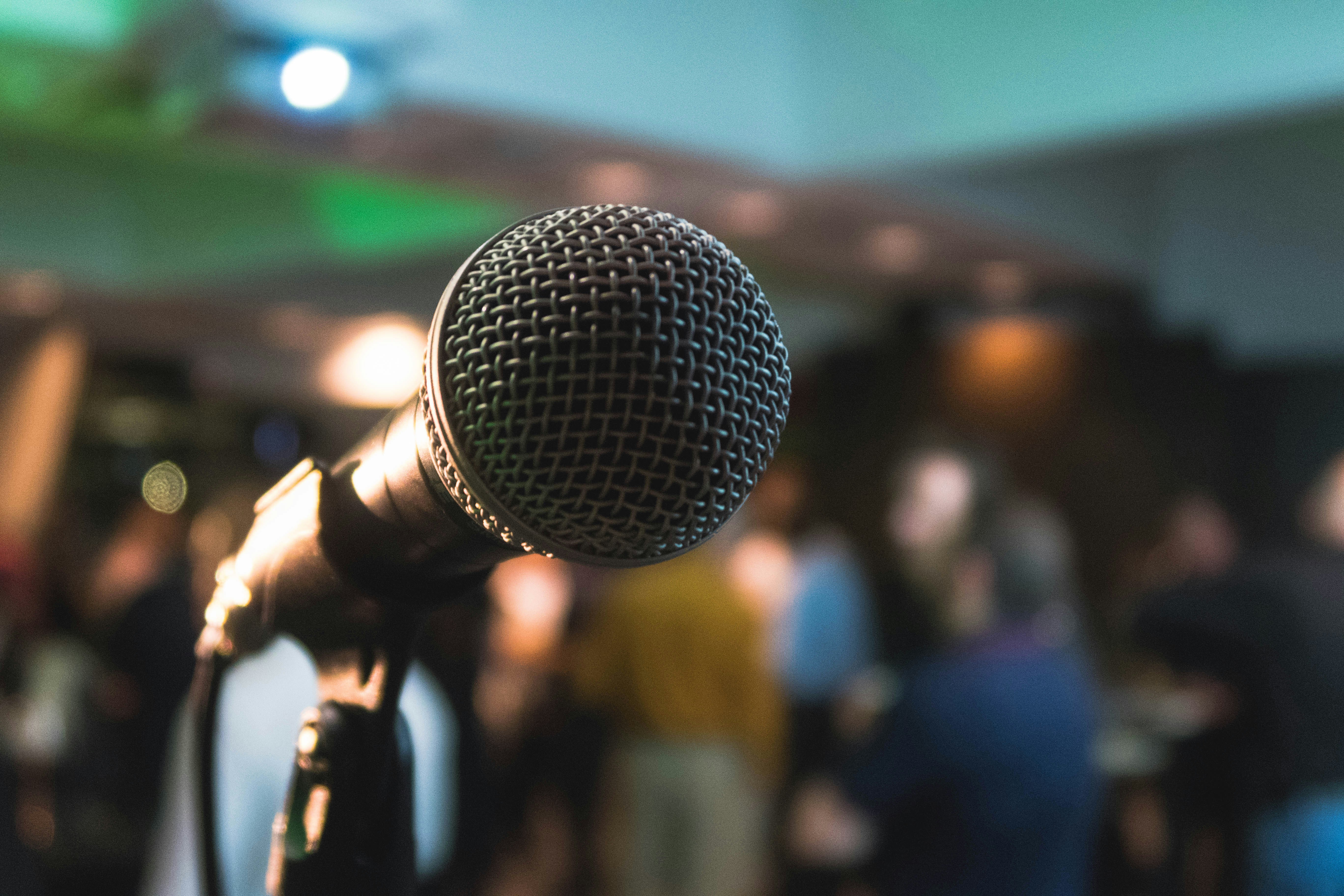 Tuvplāns mikrofona uz statīva, fonā izplūdusi auditorija ar cilvēkiem un skatuves apgaismojums.